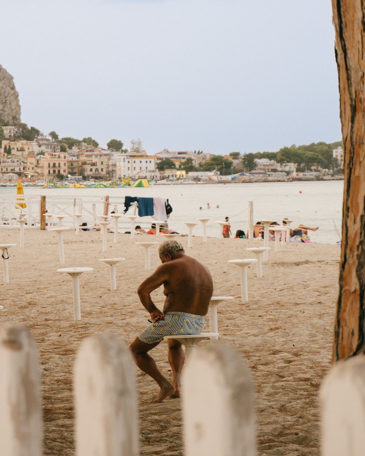 Bagnante seduto sulla battigia di una spiaggia siciliana, sullo sfondo il paese